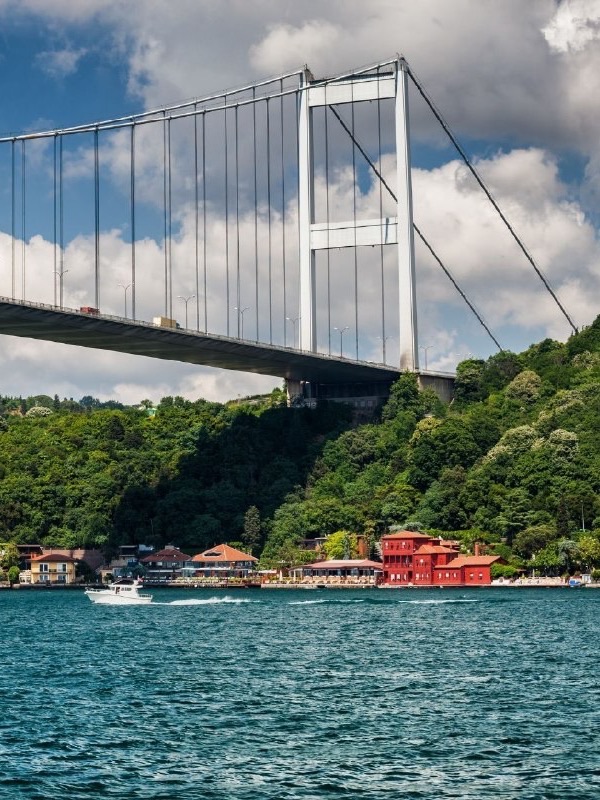 Boğaz’da Fatih Sultan Mehmet Köprüsü; altında ağaçlık yamaç, kıyıda kırmızı yalı ve açık denizde bir tekne, İstanbul